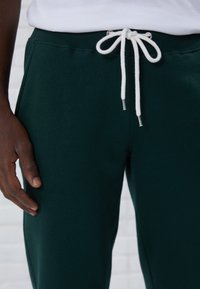 Tmavě zelené tepláky mají měkkou texturu, elastický pas a bílé šňůrky se stříbrnými koncovkami. Detailní záběr ruky na tkanině.
