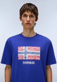 Modrá bavlněná tričko s grafikou norské vlajky s vlnitými červenými a modrými liniemi, a textem "NAPAPIJRI geographic" pod tím.