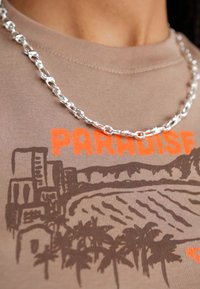Collier en chaîne argentée avec des maillons ovales entrelacés, porté sur un T-shirt beige clair avec une impression graphique d'une scène tropicale et le texte "PARADISE".