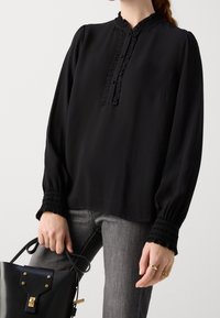 Blusa nera a maniche lunghe con tessuto texturizzato, colletto arricciato, pattina con bottoni e polsini arricciati. Abbinata a jeans grigi e a una borsa nera.