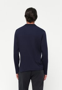 Man with dark hair wearing a navy blue long-sleeve shirt and black pants, standing facing a plain white background.