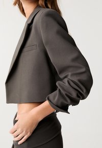 Bijgesneden donkergrijze blazer met een gestructureerde silhouet, zachte textuur, een enkele borstzak en geplooide mouwen met knoopaccenten.