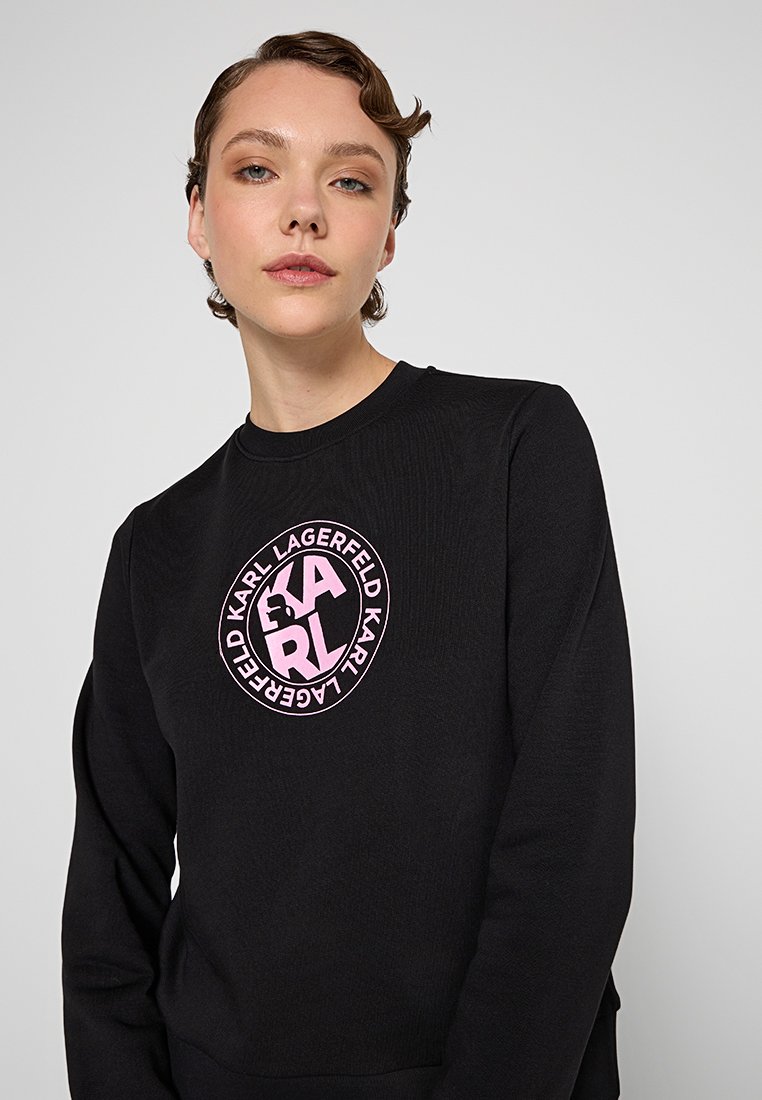 Sudadera negra con un logo gráfico redondo en color rosa, que exhibe el texto "KARL LAGERFELD" en una tipografía moderna. Mangas largas y textura de canalé.