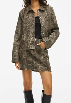 Mujer vistiendo chaqueta marrón con estampado de serpiente y minifalda a juego, con top negro y botas negras hasta la rodilla, posando con la mano en el bolsillo.