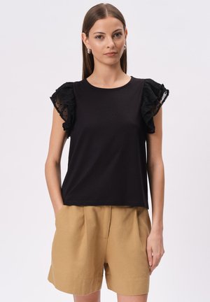 Czarna bluzka z krótkim rękawem z teksturowanymi akcentami falbanki. W połączeniu z beżowymi szortami o wysokim stanie. Gładki materiał i luźny krój.