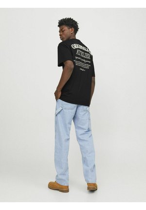 T-shirt en coton noir avec texte blanc dans le dos, associé à un pantalon cargo bleu clair. Le modèle se tient de dos, les mains dans les poches. Chaussures marron.