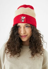 Rode en beige gebreide beanie met een ribbelstructuur, gestreept ontwerp en een geborduurd Snoopy-patch op de voorkant.