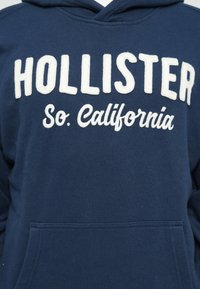 Mornarsko modra kapuca, izdelana iz mehke tkanine; vsebuje belo vezeno besedilo "HOLLISTER" in "So. California". Vključuje sprednji žep in kapuco.