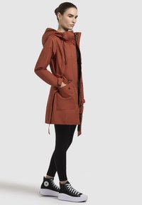 Roestrode waterdichte parka met capuchon, voorzien van trekkoorden, twee voorknooppockets en een volledige ritssluiting. Draagt over zwarte leggings en platform sneakers.