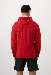 Muž otočený chrbtom, oblečený v červenej bundě s kapucňou a čiernych športových šortkách na svetlom pozadí.