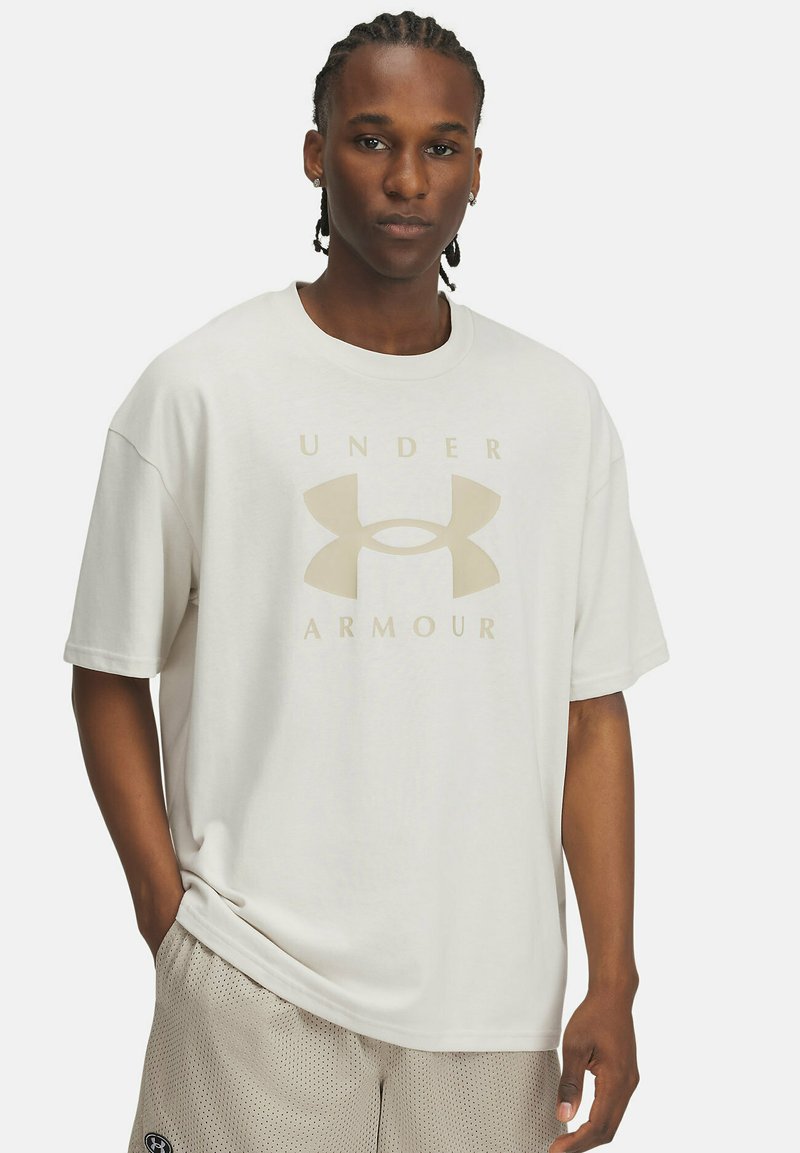 Valge puuvillane T-särk, millel on beež "UNDER ARMOUR" logo, ümar kaelus, lühikesed varrukad, mugav lõige, sile tekstuur. Kombineeritud heledate lühikeste pükstega.