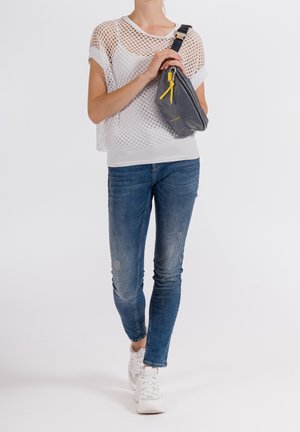 Graue Umhängetasche mit gelben Akzenten, aus glattem Material; kombiniert mit einem leichten weißen Netzoberteil und blauen Skinny-Jeans.