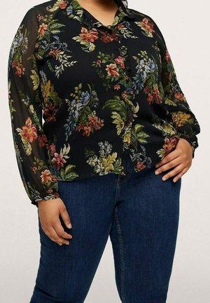 Plus-size person iført en sort bluse med knapper med farverigt blomsterprint og gennemsigtige lange ærmer, kombineret med blå jeans.