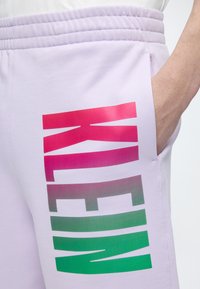 Κοντινό πλάνο σε ανοιχτό μοβ φόρμα με ελαστική μέση και μεγάλο κατακόρυφο κείμενο «KLEIN» σε κόκκινο, ροζ και πράσινο gradient στο μηρό.