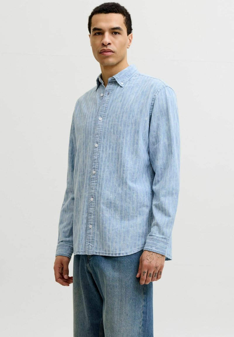 Homme debout portant une chemise bleu clair boutonnée et un jean bleu, avec des tatouages sur sa main droite, sur un fond clair uni.
