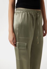 Pantaloni in satin verde oliva con vita elastica, cordoncino, tasche laterali e una tasca a toppa. Texture liscia e dettagli in metallo.