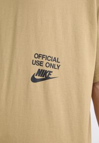 Camiseta de algodón color beige con un estampado negro que dice "USO OFICIAL SOLO" sobre el logo y el swoosh de Nike. Diseño de manga corta y cuello redondo.