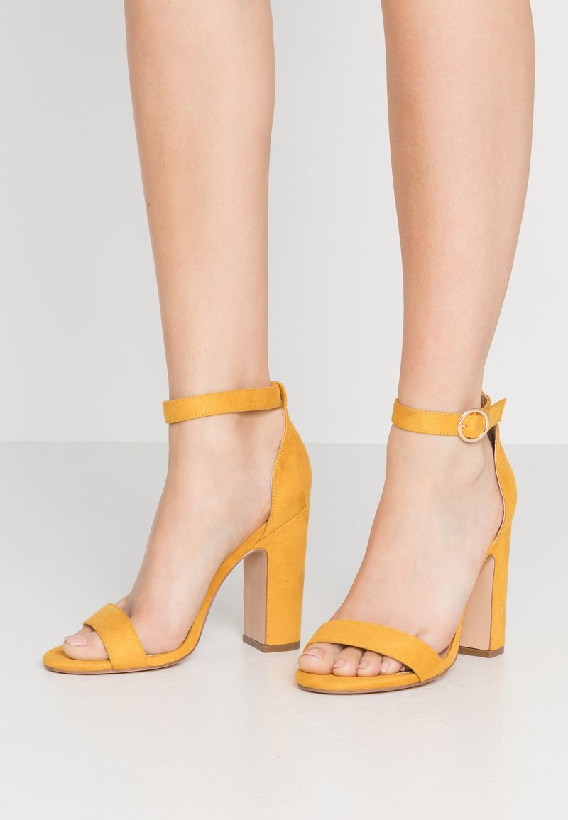 Jambes portant des sandales à talons hauts en daim jaune avec des brides cheville et des talons blocs, debout sur un sol clair devant un fond uni.