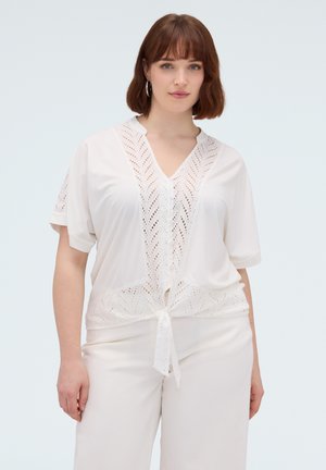 Jonge vrouw met kort bruin haar die witte broek en een witte korte mouwen blouse met kanten details en een strik aan de voorkant draagt.