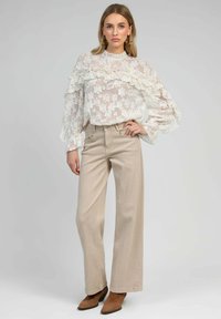 Blouse blanche en voile avec motif de dentelle florale et détails en volants, associée à un pantalon beige large et des bottes marron à bout pointu.