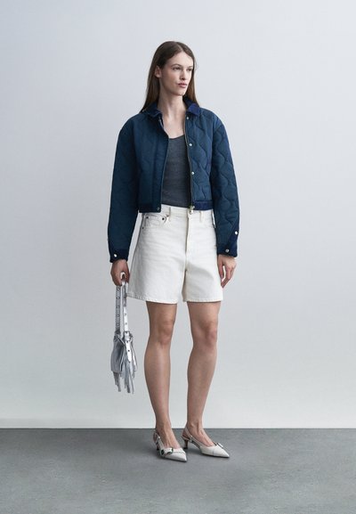 Femme portant une veste matelassée bleu marine, un débardeur gris foncé, un short blanc et des talons blancs, tenant un sac à main argenté avec des détails à franges.