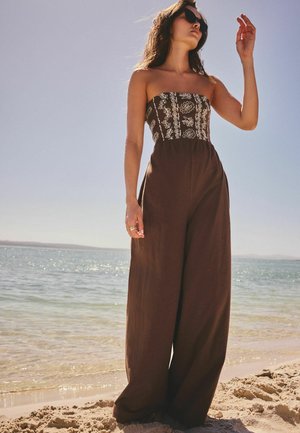 Kvinde iført brun stropløs jumpsuit med hvid broderi, solbriller, stående på sandstrand ved roligt hav under klar himmel.