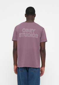 Rövid ujjú, kerek nyakú póló visszafogott lila színben, pamutból készült. A hátsó részen merész fehér "OBEY STUDIOS" felirat található. Lazább szabású.