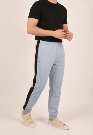 Uomo che indossa pantaloni sportivi azzurri con strisce nere laterali, sneakers beige e una maglietta nera a maniche corte, in piedi con le mani congiunte.