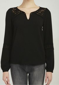Svart långärmad topp med spetsdetaljer på axlarna och ärmsluten, en V-ringning med en dekorativ nål och en avslappnad passform.