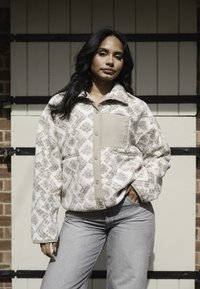 Chaqueta de forro polar en crema con un patrón geométrico, botones a presión, bolsillo sólido beige y bolsillos laterales. Llevada sobre unos jeans gris claro.