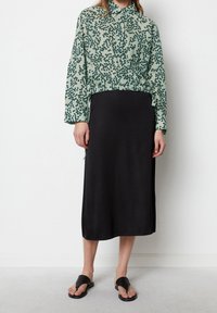 Kobieta ubrana w zielono-czarną w kratkę bluzkę z długim rękawem, czarną spódnicę midi i czarne sandały na obcasie stojąca przy białej ścianie.