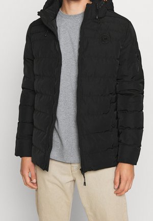 Homme portant une veste matelassée noire avec fermeture éclair devant, par-dessus un t-shirt gris et un pantalon beige, debout devant un fond uni.