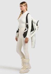 Witte lange mouwen top met een ritskraag, crèmekleurige leggings en witte sneeuwlaarzen. Een zwart-witte gewatteerde jas is over de schouder gehangen.