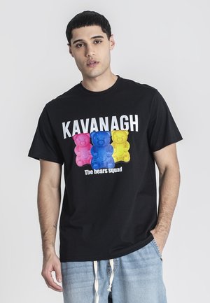 Jeune homme portant un T-shirt noir avec des oursons en gomme roses, bleus et jaunes avec le texte « KAVANAGH The bears squad », associé à un jean en denim clair.