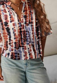 Personne portant une chemise tie-dye multicolore avec un jean bleu et un collier pendentif en forme de poisson, debout à l'intérieur devant un fond clair.
