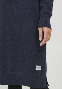 Marinblå stickad klänning i mjukt material, med långa ärmar och ribbad mudd. Har sidoöppningar och en liten vit logotags vid fållen.