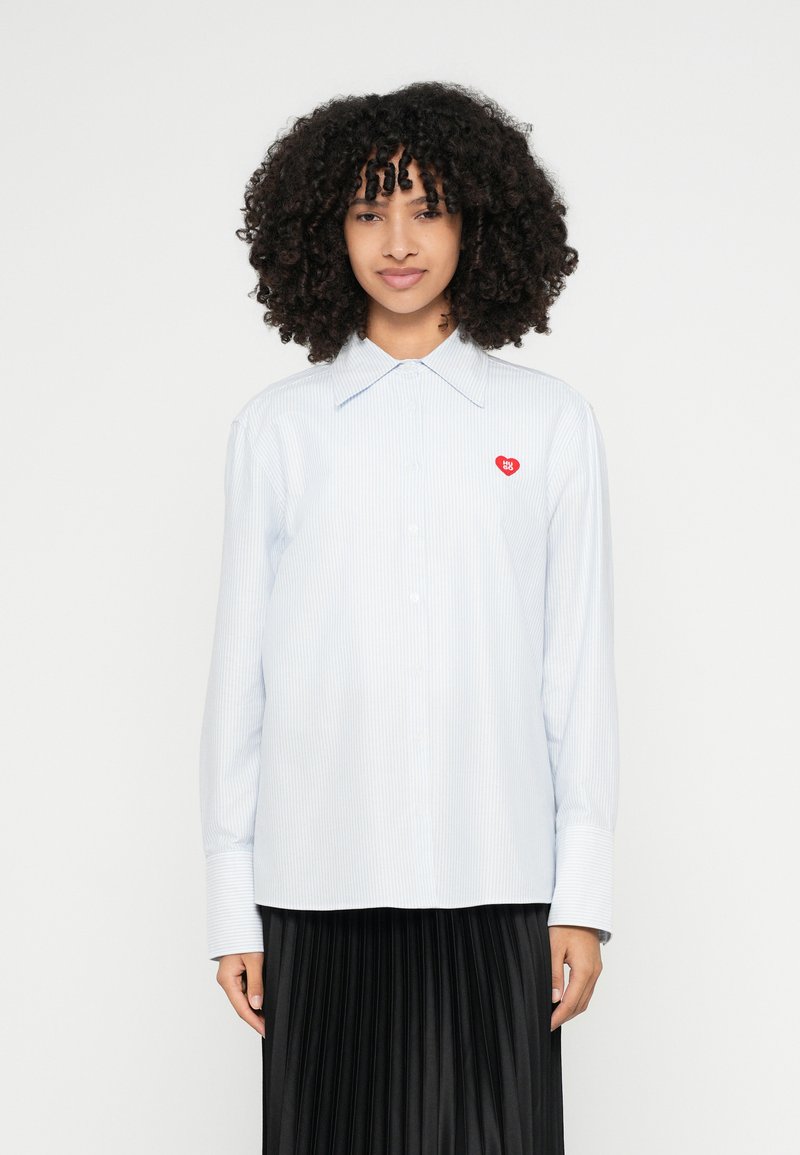 Svetlomodrá pruhovaná košeľa s špicatým golierom, dlhými rukávmi a malým červeným srdiečkom na hrudi, v kombinácii s čiernou plisovanou sukňou.