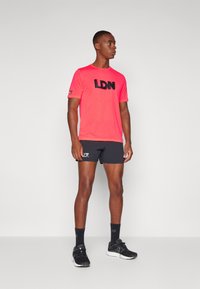 Tricou scurt roz neon cu mâneci scurte, având inscripția "LDN" în negru, asortat cu pantaloni scurți atletici negri și pantofi de alergare negri, pe un fundal simplu.