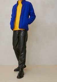 Niebieska polarowa kurtka na zamek noszona na żółtym swetrze, w połączeniu z czarnymi skórzanymi spodniami i czarnymi botkami do kostek. Gładka tekstura i nowoczesny design.