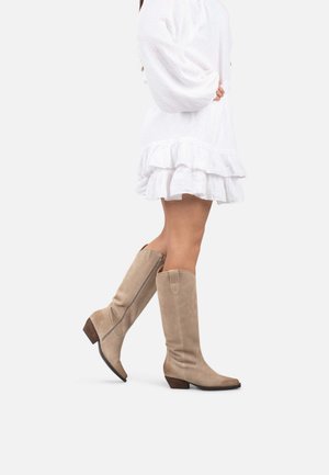 Beige Wildleder kniehohe Stiefel mit einem leichten Absatz, seitlichem Reißverschluss und glatter Textur. Getragen mit einem weißen Rüschenskleid.