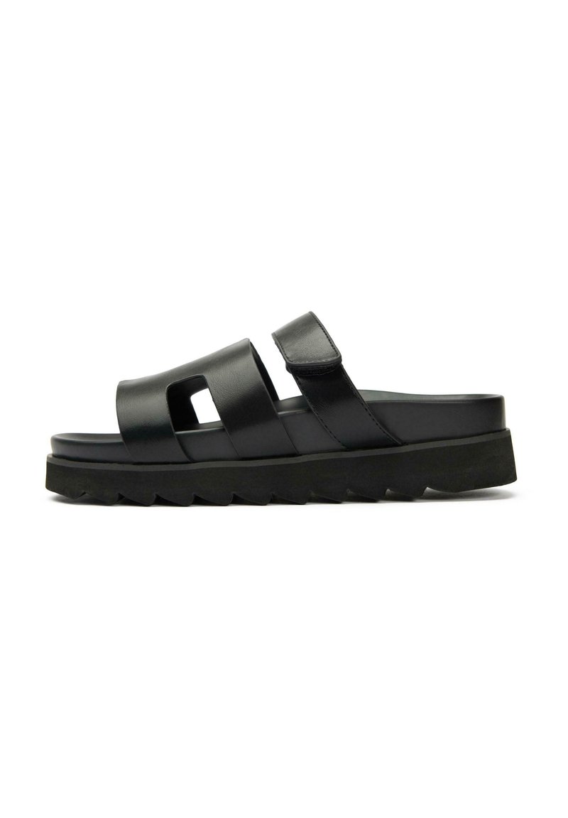 Black slide sandal with two wide straps, cut-out detail, and adjustable Velcro closure. Chunky textured sole for added grip.