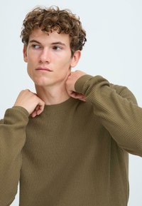 Pulover tricotat de culoare khaki, cu guler rotund, având mâneci lungi și țesătură texturată. Modelul își ajustează gulerul cu ambele mâini.