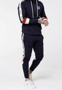 Chándal azul marino con sudadera con cremallera que presenta acentos en blanco y rojo. Los pantalones tienen franjas laterales con texto, combinados con zapatillas blancas.