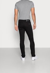 Man som bär svarta slim-fit jeans, grå t-shirt och vita sneakers, står vänd bort från kameran mot en helt vit bakgrund.