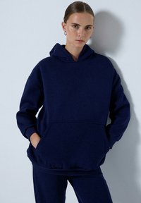Námořnická modrá oversized mikina z měkké látky, s přední kapsou a žebrovanými manžetami, nošena s ladícími kalhotami.