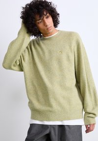 Maglione verde chiaro in un morbido tessuto a maglia testurizzata, caratterizzato da una vestibilità comoda, polsini a costine e un piccolo logo sul petto. Indossato sopra una camicia bianca.