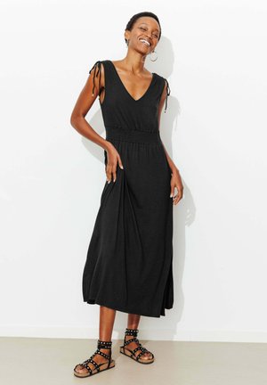 Femme souriante debout contre un mur blanc, portant une robe midi noire sans manches avec des bretelles nouées aux épaules et des sandales plates noires cloutées.