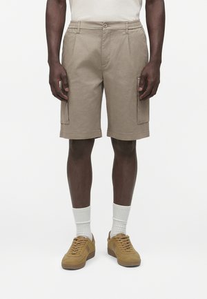 Mann trägt beige Cargo-Shorts, weiße Sportsocken und senfgelbe Sneaker und steht vor einem einfarbigen Hintergrund.