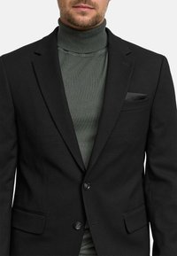 Svart strukturert blazer med notched lapels og to knapper, brukt over en tettsittende mørkegrønn turtleneck. Lommerekke som aksent på blazeren.