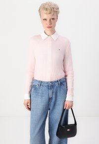 Suéter de punto rosa claro con un collar blanco, combinado con unos jeans de mezclilla azul de cintura alta y un pequeño bolso negro con un logotipo.